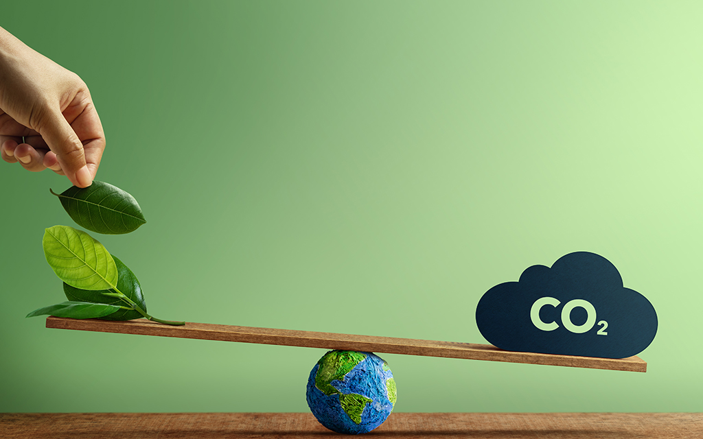 Hand placing green leaves on a balance scale with the Earth and a CO2 cloud, representing the balance between renewable carbon and emissions.