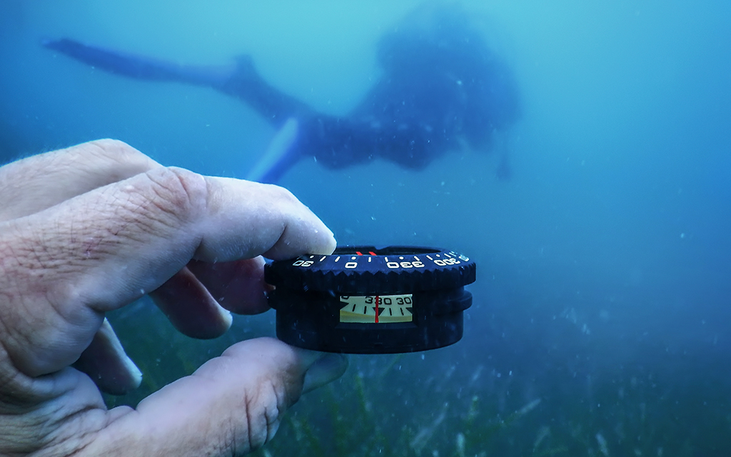Diver navigating underwater using a compass, with a scuba diver visible in the background