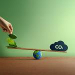 Hand placing green leaves on a balance scale with the Earth and a CO2 cloud, representing the balance between renewable carbon and emissions.