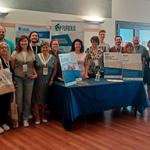 A group of people standing and posing at a scientific conference. In front of them, in the centre, is a table displaying the posters and prototypes that were shown at the conference.